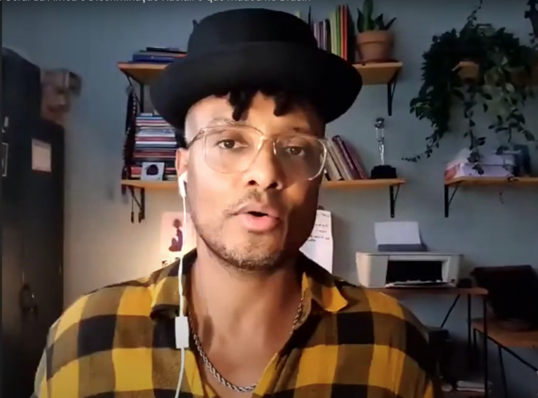 Print de uma tela mostrando um homem negro de chapéu preto, óculos de grau e camisa xadrez com tons de amarelo e preto