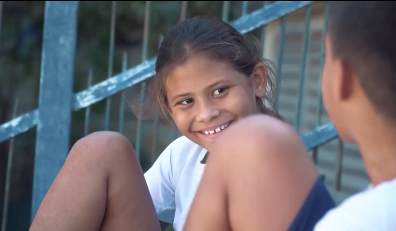 Menina morena está sorrindo e interagindo com outra criança, que está de costas para quem bateu a foto