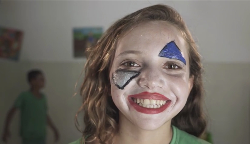 Rosto de uma menina branca e loira pintado com maquiagem similar a de um palhaço. Ela está sorridente demais
