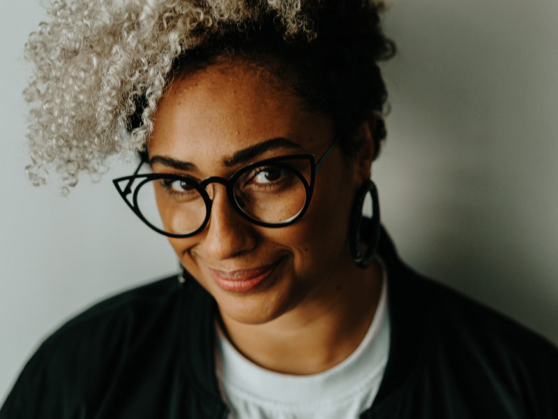 Uma jovem mulher negra de cabelos loiros encaracolados, óculos de grau de armação preta e um leve sorriso sem mostrar os dentes