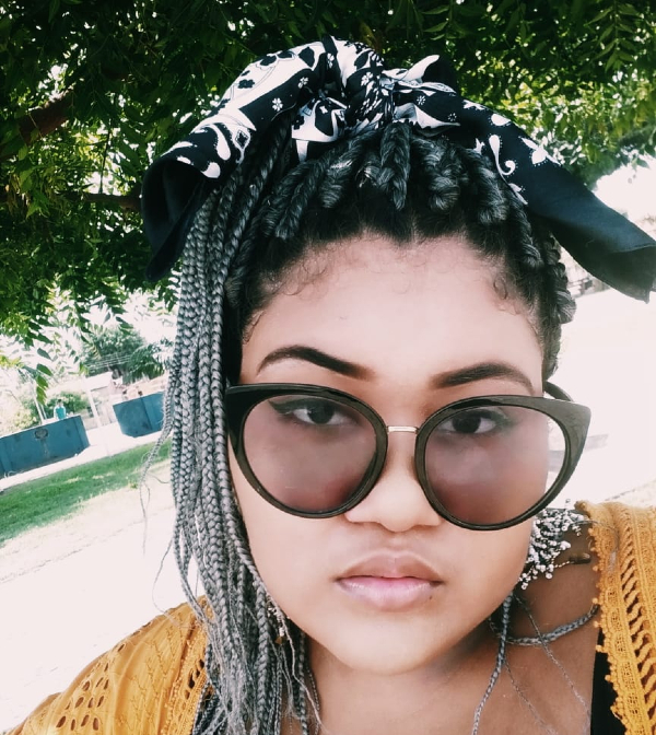 Foto de Dandara, que na imagem é uma jovem mulher negra com tranças no cabelo comprido. Ela está usando um óculos de sol de armação redonda
