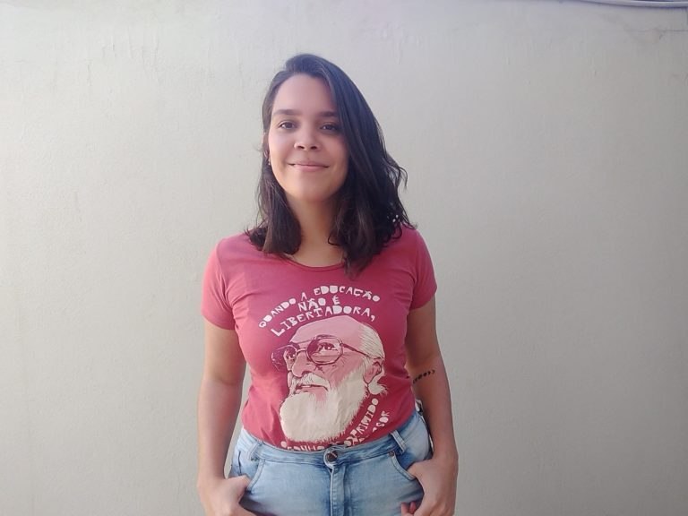 Imagem de Daniele, que na foto é uma menina branca de cabelos médios e está com as mãos nos bolsos de sua calça jeans e camisa vermelha