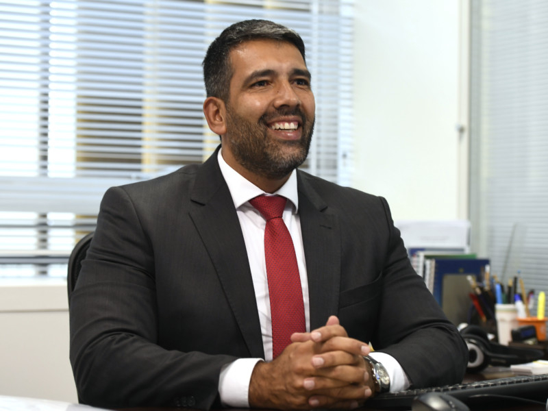 Imagem de um homem moreno de barba ralinha bem aparada, cabelo curto bem tratado e sorridente. Ele está trajando um terno, com a gravata vermelha