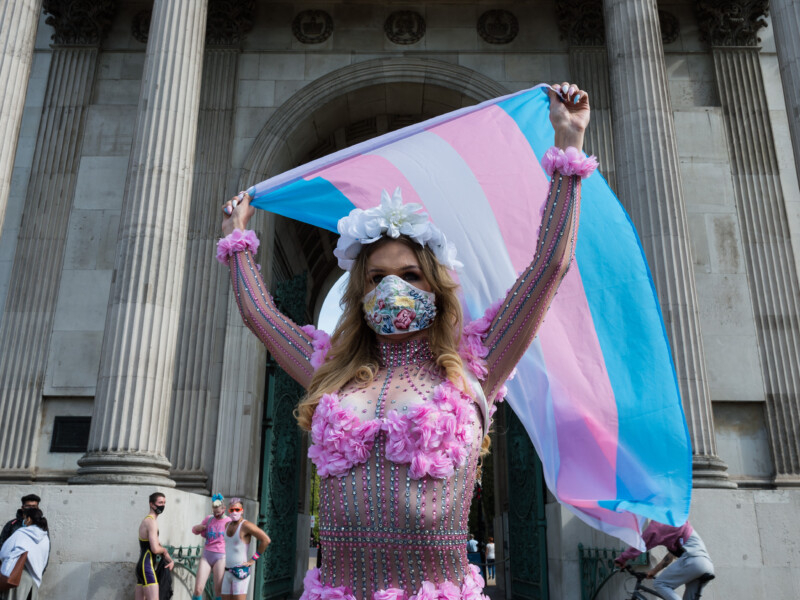 Uma mulher branca transexual está de máscara de prevenção à covid-19 e levantando uma bandeira do orgulho transgênero