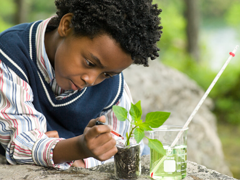 Um menino negro está mexendo em um pequeno vasinho de planta. Parece que ele está pingando algum líquido na terra onde a plantinha está inserida