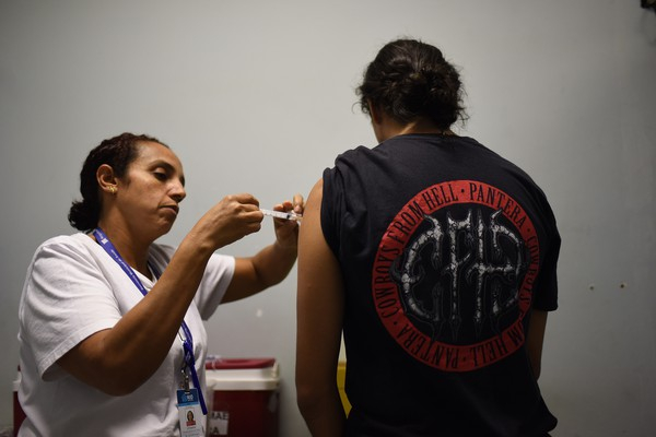Uma pessoas de costas para quem bateu a foto está sendo vacinada no braço esquerdo por uma enfermeira