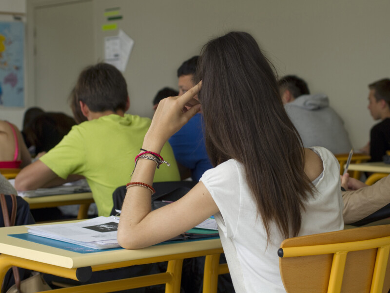 Alunos sentados em sala de aula. A foto foi batida no fundo da sala, então estão todos de costas para a imagem