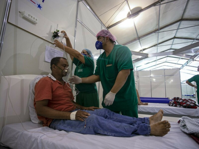 Um homem está sentado em uma cama de hospital, respirando com a ajuda da máscara de oxigênio e sendo atendido por dois enfermeiros