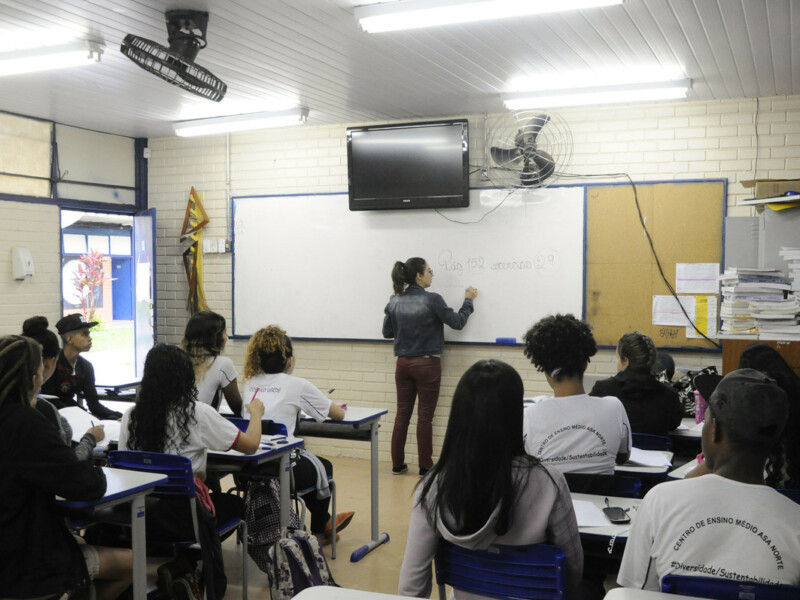 Uma sala de aula com a professora escrevendo algo no quadro ao fundo e os alunos sentados observando