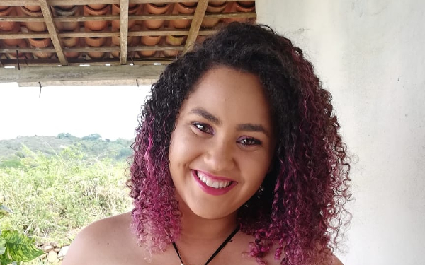 Foto de Felícia, uma jovem negra de cabelos crespos com tons de cor de rosa. Ela está sorridente e maqueada