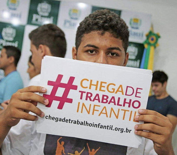 Foto de Felipe, que na imagem é um jovem rapaz negro de cabeço curto e encaracolado. Ele está segurando um cartaz que esconde a metade de baixo do rosto. O cartas tem os dizeres: #ChegaDeTrabalhoInfantil e o site da ONG