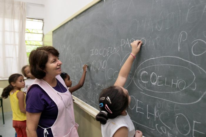 Uma professora está olhando uma aluna pequenina escrevendo no quadro negro em sala de aula