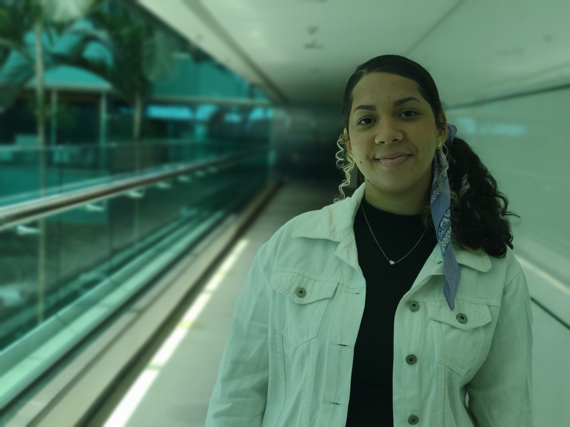 Uma jovem sorri. Ela estrá com o cabelo preso em um rabo de cavalo e usa um casaco jeans claro. Ao fundo, o corredor de um prédio com placas de vidro.