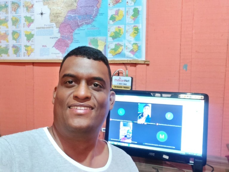 Um homem negro, sorridente, de costas para uma tela de computador e de frente para quem bateu a foto