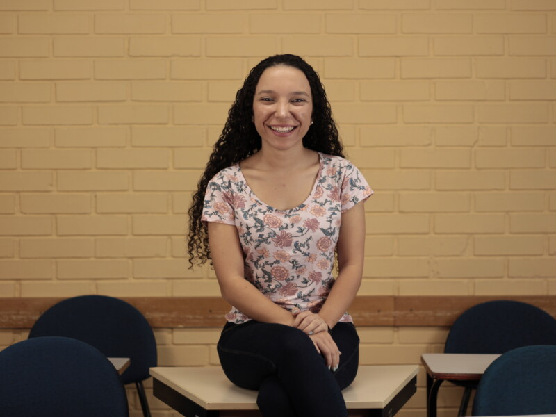 Uma mulher branca está sentada de pernas cruzadas olhando sorridente para quem bateu a foto. Ela tem cabelo cacheado e comprido e está de calça jeans escura e uma blusa com estampas coloridas