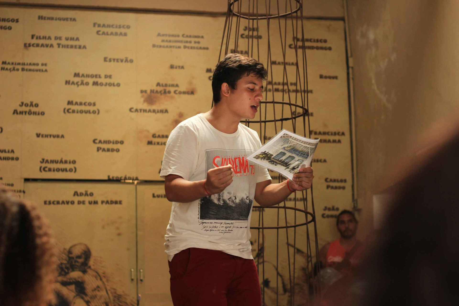 Imagem de um homem em pé, trajando uma bermuda cor vinho e uma camisa branca. Ele parece estar recitando uma poesia em público