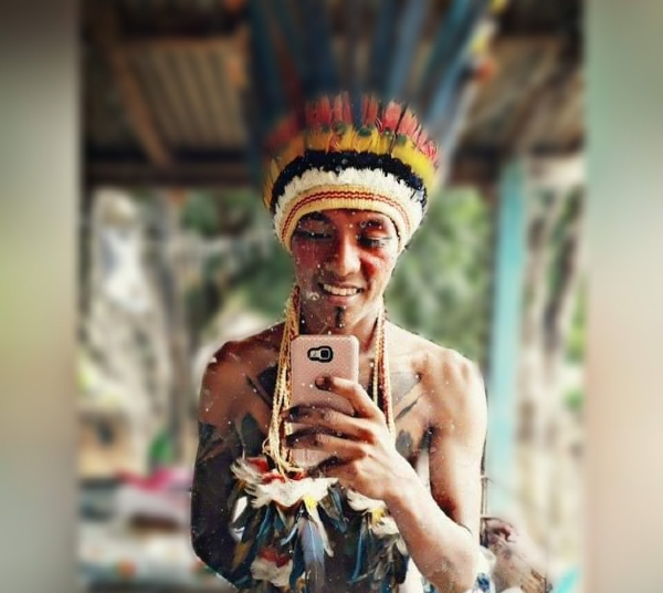 Foto de Jeseias, que na foto é um rapaz magro e trajado com coquá indígena. Ele parece estar fazendo uma foto em frente a um espelho com seu celular cor de rosa
