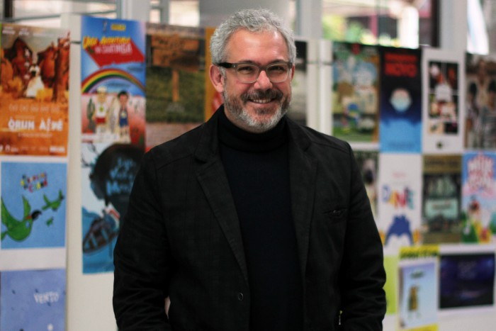 Um homem sorridente, de barba e cabelo branco, de óculos de grau e vestindo camisa preta e um blazer preto por cima