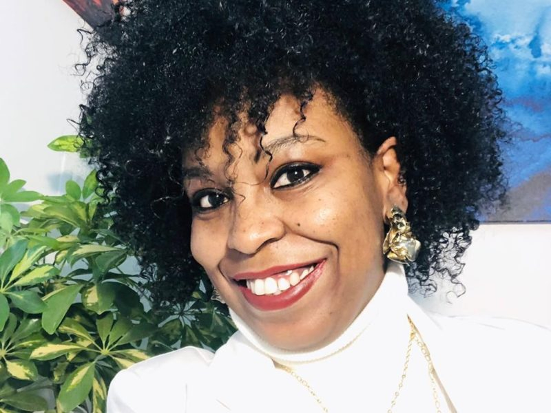 Foto de Joice, uma mulher negra de cabelo volumoso e encaracolado. Ela está de brincos dourados, batom vermelho e uma camisa branca de gola rolê