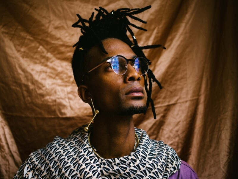 Homem magro, negro, de óculos de grau, parece estar usando um cachecol nas cores preta e branca, usa um brinco dourado com design semelhante a um microfone e tem cabelo escuro com vários dreads de tamanhos diferentes
