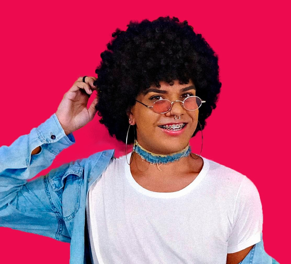 Imagem de Kevyn, uma pessoa negra de cabelos volumosos encaracolados e bem pretos. Está usando cordão azul, uma blusa branca, aparelho nos dentes, piercing no nariz e óculos estiloso