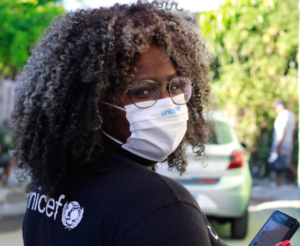 Foto de Lays, que na imagem é uma jovem mulher negra de cabelos volumosos, mais claros e encaracolados. Ela está usando óculos de armação redonda, camisa da UNICEF e máscara de prevenção à covid-19