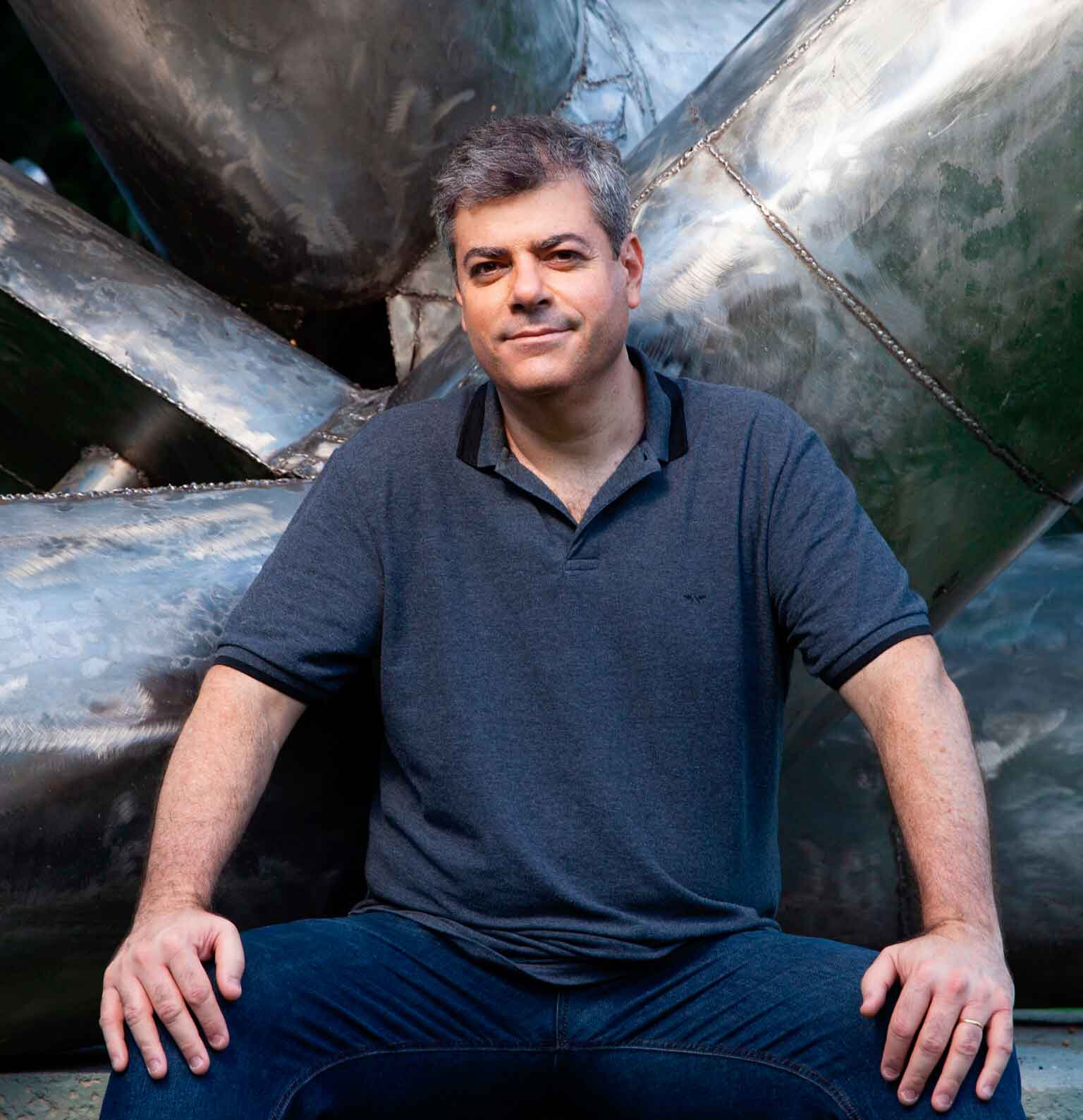 Imagem de um homem branco, de cabelos semi grisalhos, sentado e trajando uma calça jeans e camisa pólo