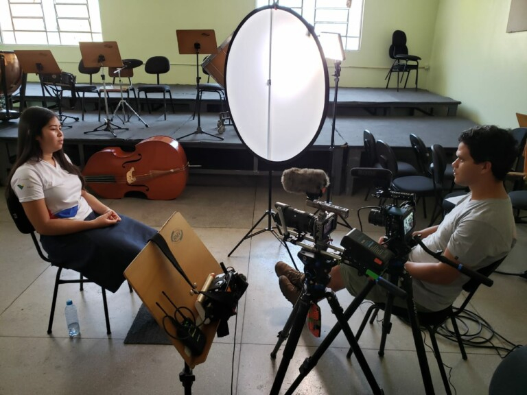 Uma menina de uniforme escolar está sentada diante de um homem com uma máquina de filmar presa a um tripé. Ela parece estar dando entrevista ao rapaz