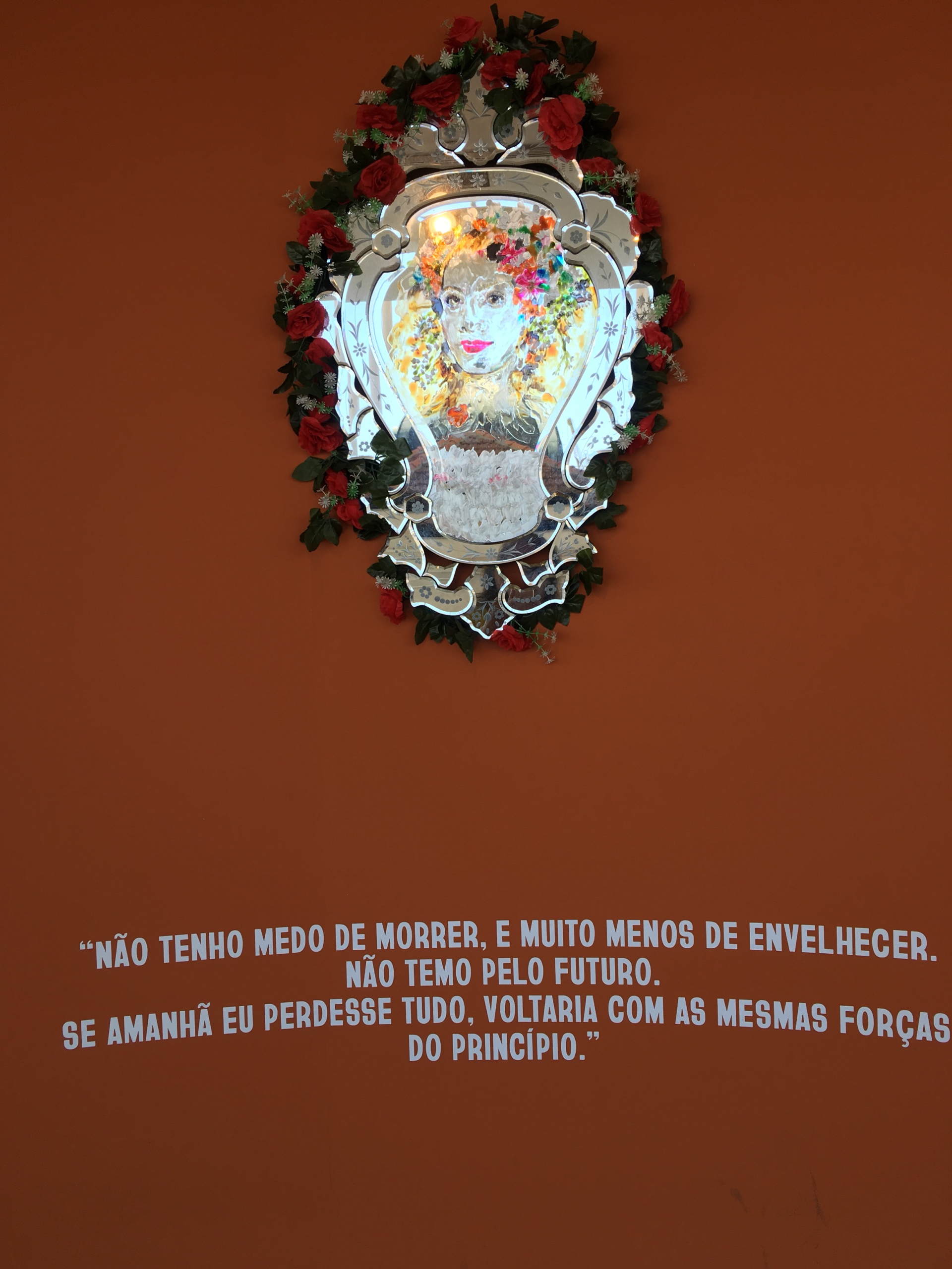 Sobre um fundo marrom, uma arte espelhada (sobre uma armação entalhada) mostra o rosto de uma mulher.