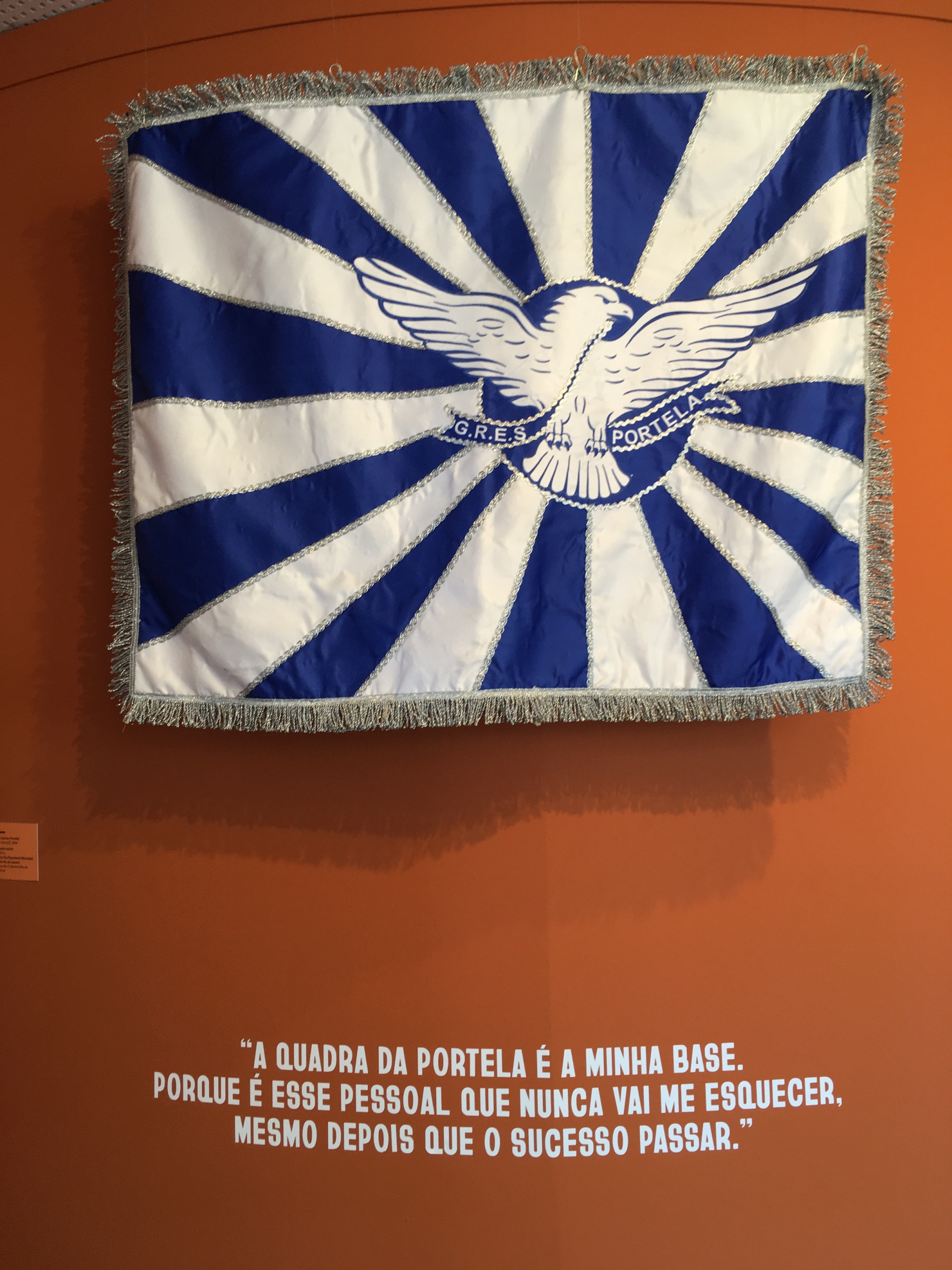Sobre um fundo marrom, a bandeira da escola de samba Portela, em faixas azuis e brancas que saem do centro da imagem, onde está uma águia.
