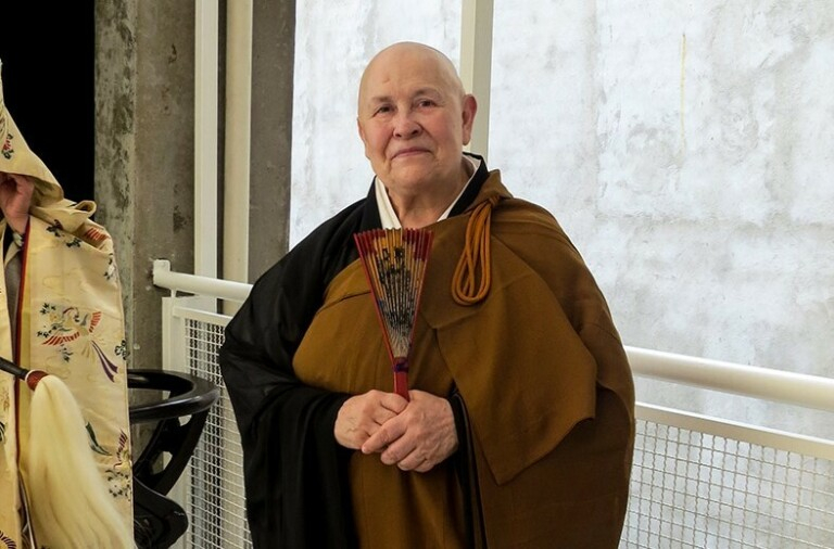 Mulher careca olhando seriamente para quem bateu a foto. Está trajando roupa característica dos monges e um leque fechado está em suas mãos