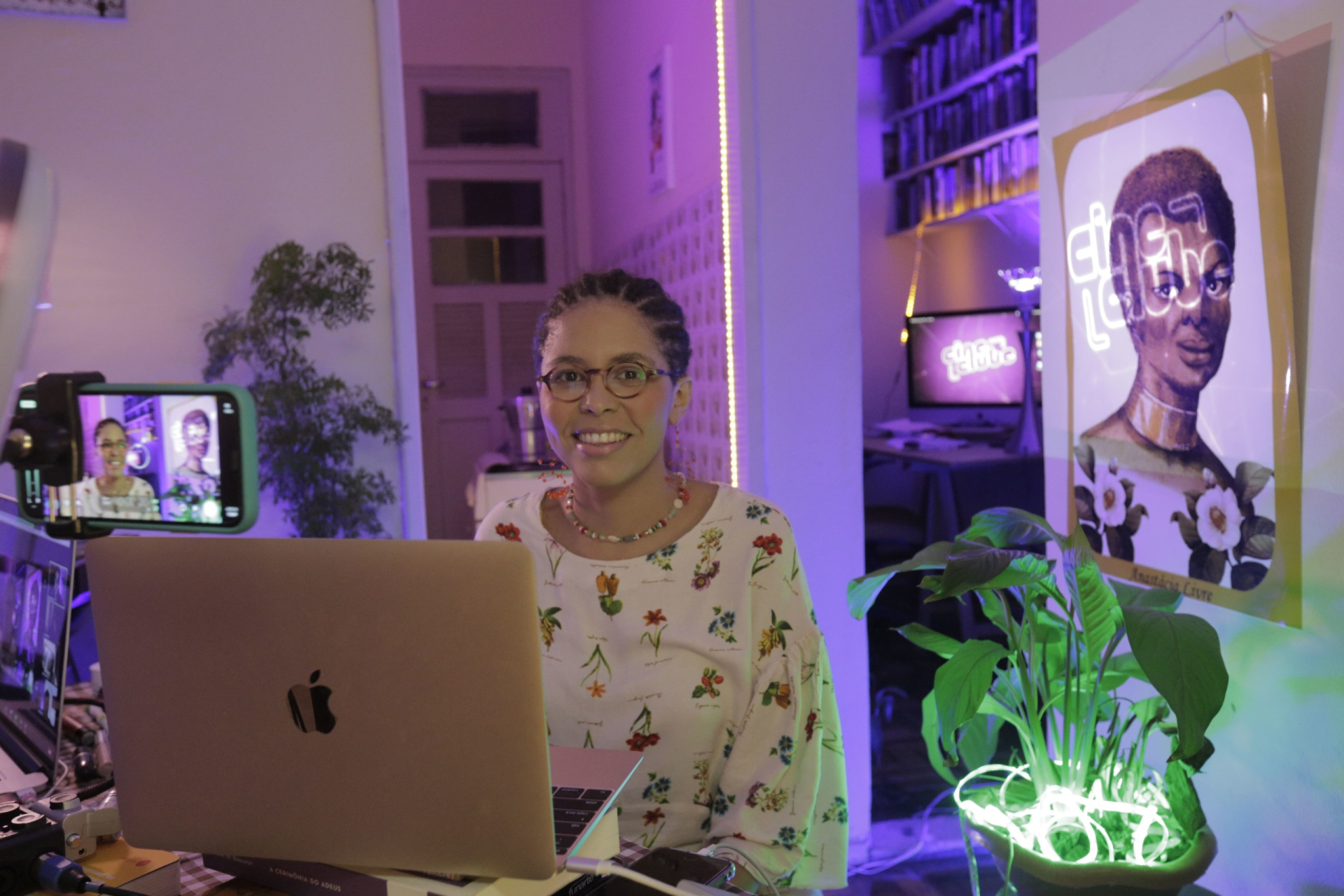 Imagem de uma mulher negra, de cabelos bem curtos e óculos de grau. Ela está sorridente no centro da imagem, com um notebook aberto à sua frente e o cenário do programa Cineclube Futura ao fundo