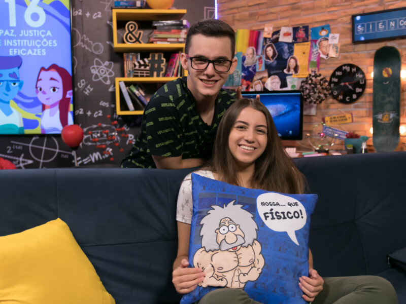 Um rapaz e uma menina jovens, brancos e sorridentes para quem fez a foto. Estão em um estúdio de TV, com o cenário do programa ao fundo, repleto de elementos coloridos e científicos modernos. A menina está sentada em um sofá segurando uma almofada azul de Einstein e o menino está em pé atrás do sofá