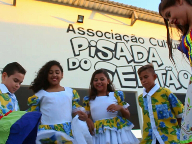 Quatro crianças e uma mulher adulta estão lado a lado trajando roupas típicas de dança, bem coloridas com as cores do Brasil