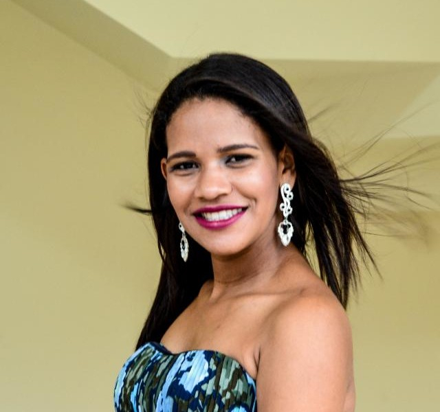 Imagem da professora Priscila, uma jovem mulher negra, de cabelos escuros e alisados, brincos prateados compridos. Ela está bem arrumada e maquiada na foto.