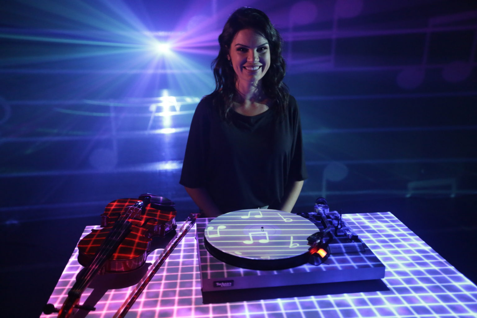 Uma mulher sorridente está em pé e atrás de uma mesa, que contém um violino e uma vitrola. Há uma luz brilhante ao fundo e parece haver uma espécie de globo de luz, pois a imagem está toda cheia de pequenas sombras de notas musicais