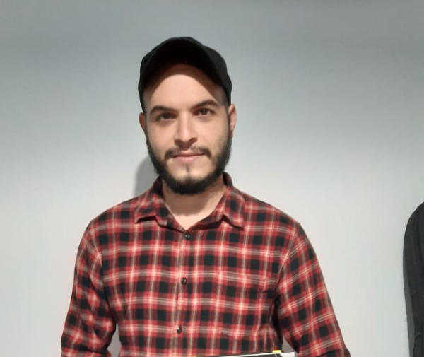 Foto de Raul, que na imagem é um jovem branco de barba bem aparada. Ele está usando camisa xadrez vermelha com tons de preto e branco e um boné preto
