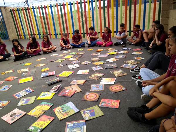 Foto de crianças uniformizadas em um pátio de colégio. Elas estão todas sentadas formando um círculo e, dentro da circunferência, há dezenas de livros coloridos amontoados
