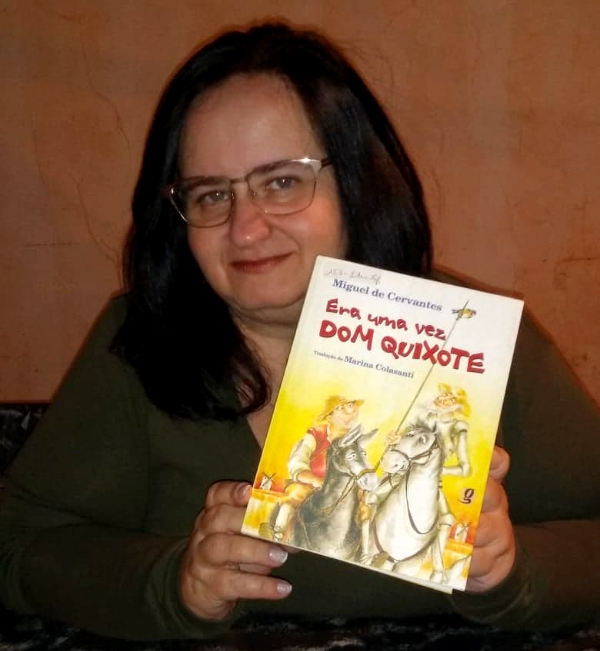 Foto de Rita, uma mulher branca na casa dos 40 anos de óculos de grau e cabelos pretos. Ela está posando para a foto segurando seu livro favorito, a obra "Era uma vez Dom Quixote"