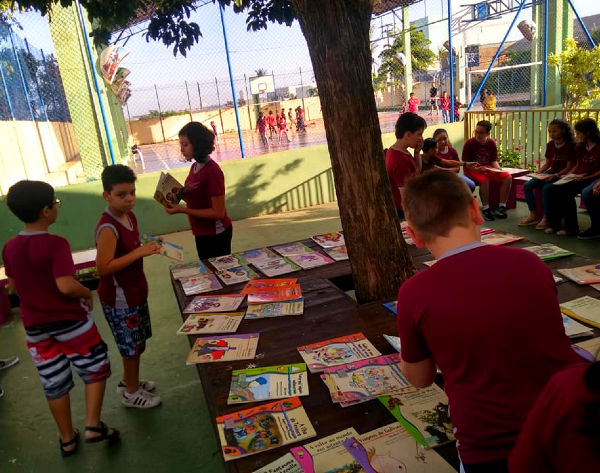 Imagem de dezenas de livros amontoados ao redor de uma árvore plantada em um pátio de colégio. Há vários alunos uniformizados pegando os livros, outros já parados e lendo