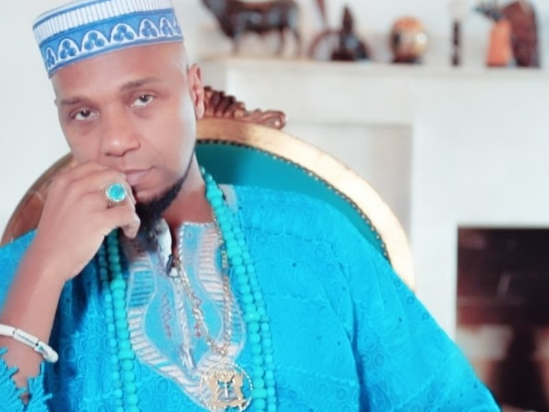 Um homem negro com roupas características de cultura africana está olhando sério para quem bateu a foto e com a mão direita encostando em sua própria boca e bochecha