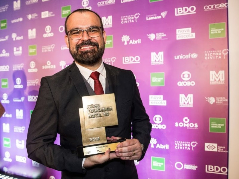 Imagem de Rodrigo, um homem branco, barbudo, quase totalmente careca, de óculos de grau, terno, gravata vermelha e está sorrindo e segurando um troféu de Educador Nota 10