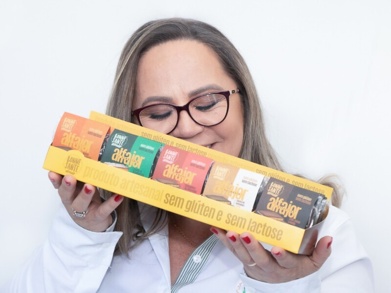 Mulher branca, de óculos de grau e cabelos soltos está segurando um objeto comprido amarelo, que contém cinco embalagens de alfajor