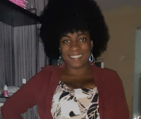 Foto de Taciane, uma jovem mulher negra de cabelo volumoso no estilo "black power" e brincos de argola. Ela está sorridente e trajando uma blusa vermelha aberta por cima de um top branco
