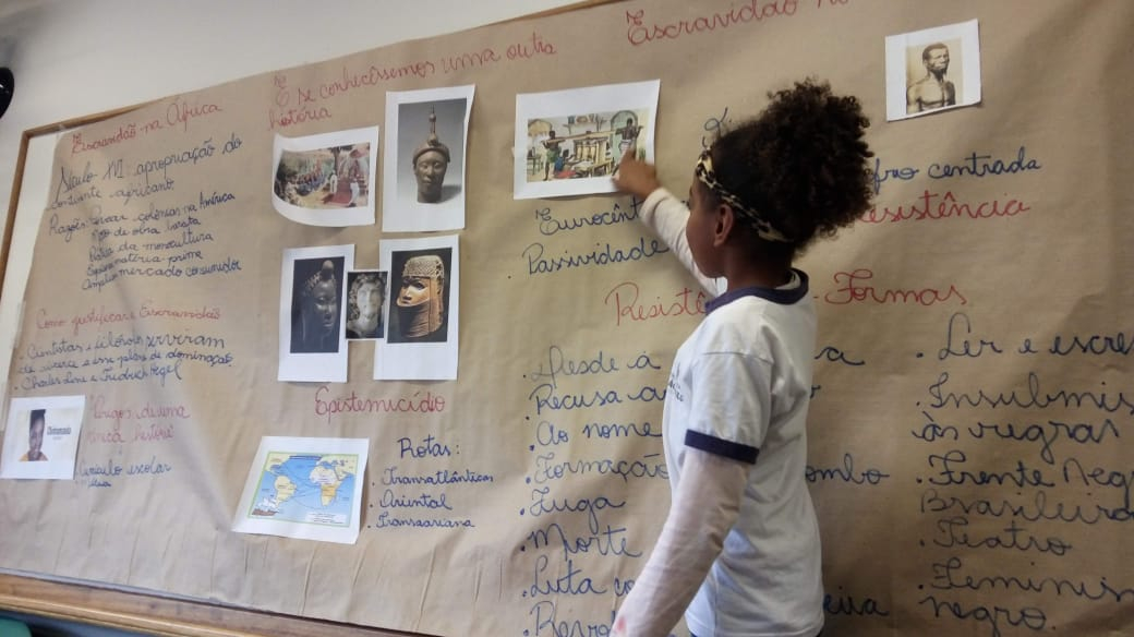 Uma estudante negra, criança, está em pé em frente a um mural apontando para uma foto neste. É um mural com história afro-brasileira