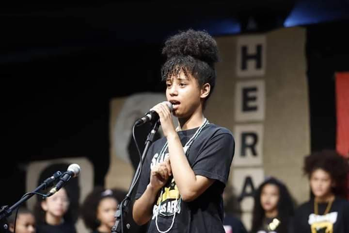 Uma adolescente negra está em pé, segurando um microfone com a mão esquerda e parece estar falando no momento da foto. Ao fundo, há pessoas sentadas observando a menina