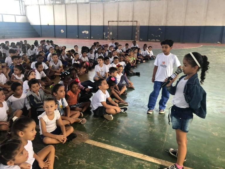 Uma menina está em pé em um ginásio de escola segurando um microfone com a mão direita e falando com outras dezenas de crianças, que estão sentadas à sua frente, espalhadas pelo chão do ginásio