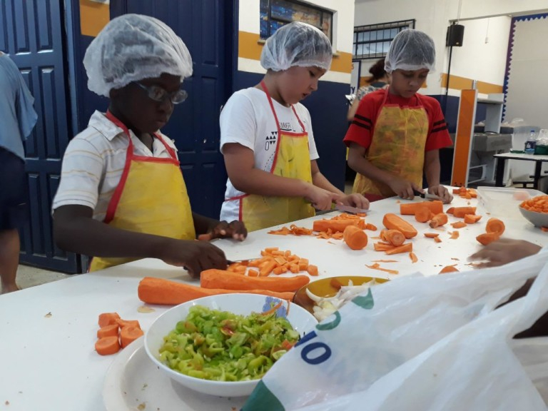 Três crianças estão lado a lado cortando cenouras em uma grande mesa de cozinha. As crianças estão com toucas de cozinha e aventais amarelos