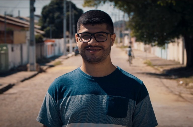 Um homem moreno, de óculos e camisa azul está posando para a foto no meio da rua