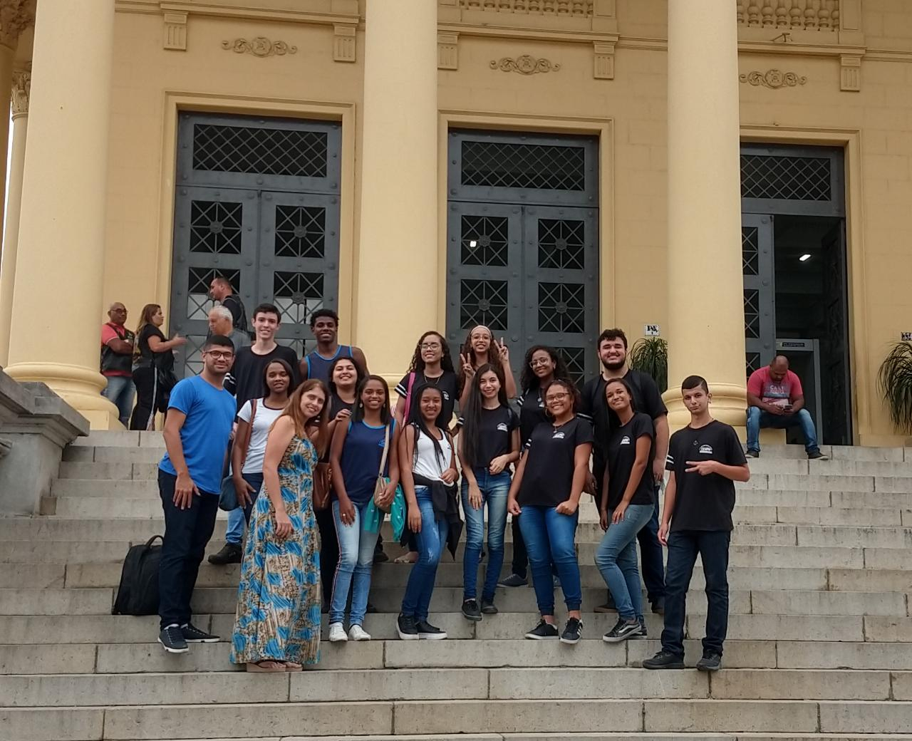Estudantes ao lado do professor em uma fachada de escola possivelmente com escada de concreto